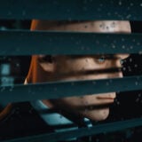 Agent 47 peers through window blinders at his target.